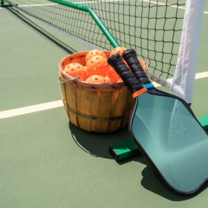 Two pickleball balls and a paddle lying on a green court surface with white boundary lines. The paddle is green and black with a perforated face and a black grip handle.
